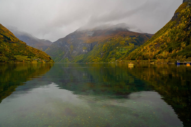 Shoreline of a cloud covered fjord surrounded by steep mountains and vegetation in yellow and orange fall colors. . High quality photo - Fotografie, Obrázek