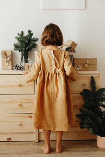 Christmas Advent calendar for kids. Girl wearing mustard linen dress looking at basket with advent gifts boxes on wooden cabinet. Eco friendly christmas - Fotoğraf, Görsel