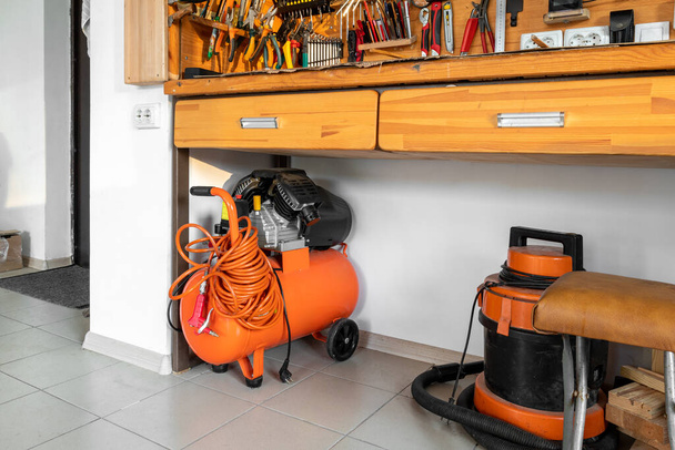 Orange kleine tragbare industrielle Power-Luft-Kompressor mit Spulenschlauch und Druckluftpistole und Staubsauger zu Hause Lagerhaus-Garage unter Holzwerkbank. Elektrische Werkzeuge und Geräte. - Foto, Bild