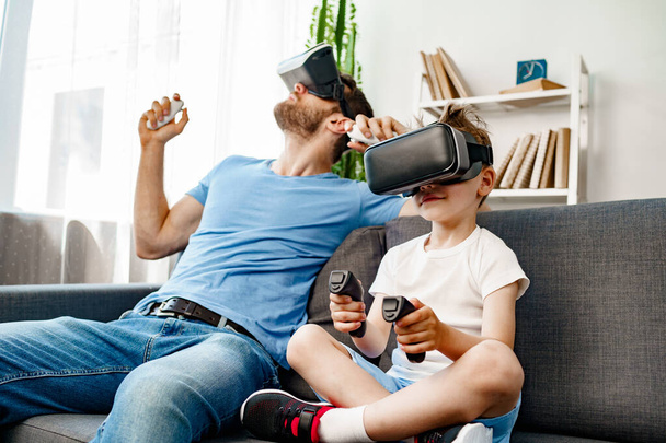 Dad and son sitting on sofa and playing games with VR glasses - Foto, imagen