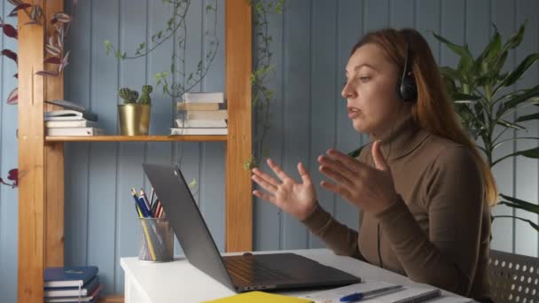 Side view, young woman discuss common project with colleague by video conference connection, sit a desk wear headphones talks with classmate studying together using video call application and laptop - Video, Çekim