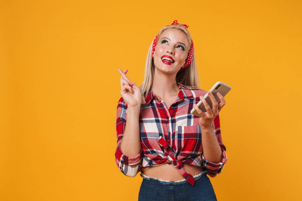 Εικόνα χαρούμενη ξανθιά pinup κορίτσι κρατώντας τα δάχτυλα σταυρωμένα, ενώ χρησιμοποιώντας κινητό τηλέφωνο απομονώνονται σε κίτρινο φόντο - Φωτογραφία, εικόνα