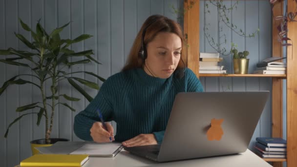 Μαθήτρια κάθεται στο γραφείο φορούν ακουστικά ματιά στην οθόνη του υπολογιστή σημειώνοντας στο βιβλίο άσκηση μελέτη προετοιμάσει τη χρήση δοκίμιο on-line σύνδεση στο διαδίκτυο, πληροφορίες αναζήτησης, αυτο-εκπαίδευση έννοια - Πλάνα, βίντεο
