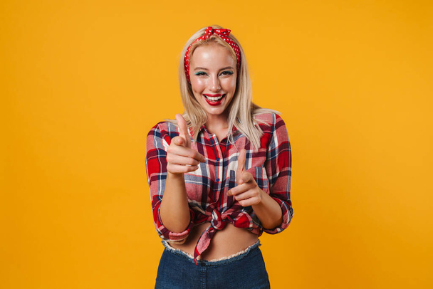 Image of cheerful beautiful pinup girl smiling and pointing fingers at camera isolated over yellow background - Foto, Imagen