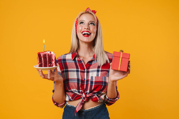 Image of happy charming pinup girl posing with gift box and birthday cake isolated over yellow background - 写真・画像
