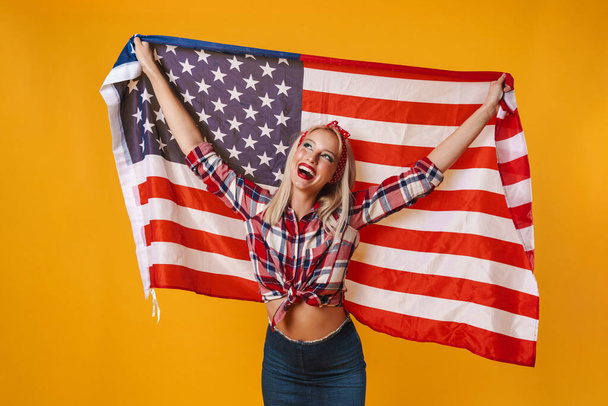 Image of happy charming pinup girl posing with american flag and smiling isolated over yellow background - Foto, Imagen