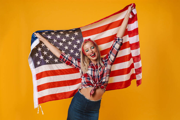 Image of happy charming pinup girl posing with american flag and smiling isolated over yellow background - Photo, Image
