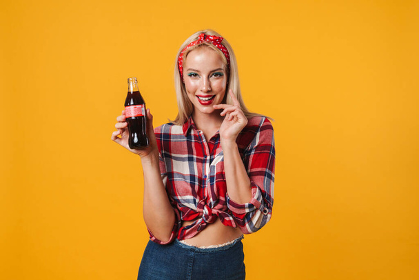 Image of happy charming pinup girl posing with soda and smiling isolated over yellow background - Photo, Image