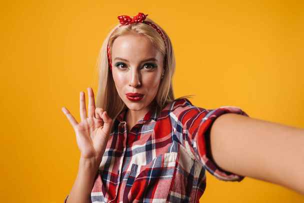 Image of cheerful pinup girl showing ok sign while taking selfie photo isolated over yellow background - Fotoğraf, Görsel