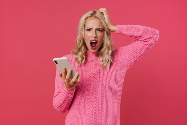 Image of furious blonde girl screaming while holding mobile phone isolated over pink background - 写真・画像
