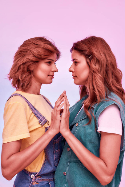 Portrait of two attractive young girls, twin sisters looking at each other, joining palms, posing together isolated over pink background - Φωτογραφία, εικόνα