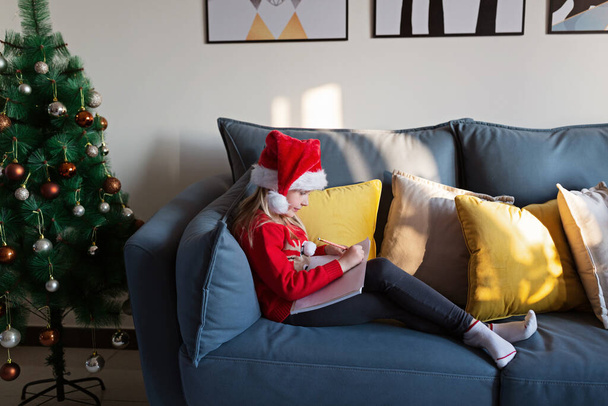 Cute blonde caucasian girl 7 years old sitting on couch near Christmas tree and writing letter to Santa Claus. Bedroom decorated with garlands. Xmas celebrate, happy new year 2021. Time to make wishes - Φωτογραφία, εικόνα