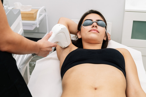 Ästhetikerin bei der Behandlung einer Frau mit Laserdioden in der Achselhöhle, wo die Kundin eine Laserschutzbrille trägt - Foto, Bild