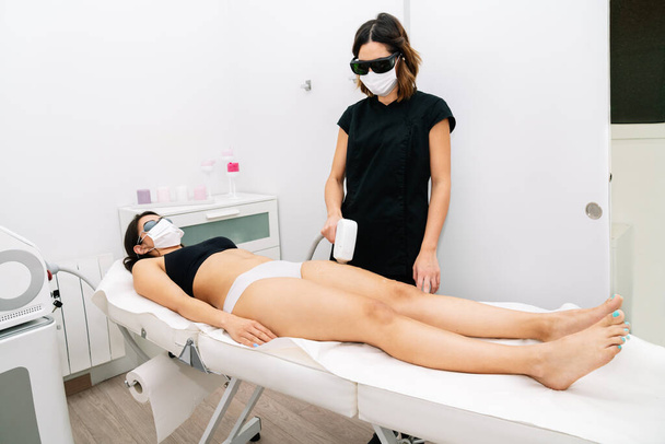 太ももの女性にレーザー脱毛の治療を与え、 2020 covid19コロナウイルスパンデミックのために顔マスクを身に着けているエステティシャン - 写真・画像