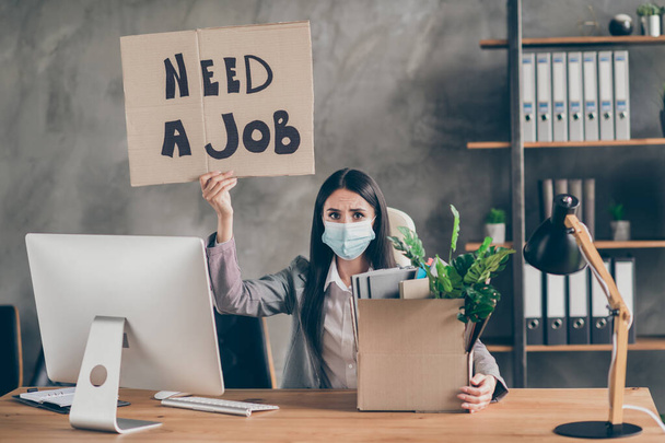 Photo of frustrated sad upset girl with medical mask sit desk table lost job corona virus quarantine company bankrupt crisis hold cardboard text box in workplace workstation - Photo, Image