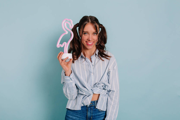 Studio photo of an inspired lady posing with smile holding pink flamingo over blue background. Indoor portrait of relaxed young girl - Fotoğraf, Görsel