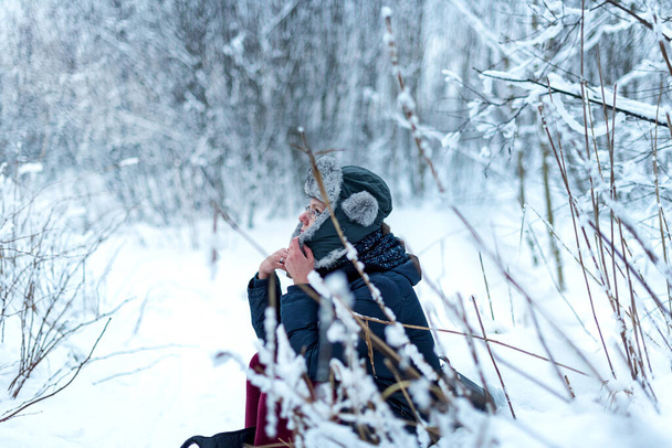 Una donna siede tra gli alberi innevati in una foresta invernale e gode della prima neve. Con un cappello con paraorecchie. Il concetto della vacanza invernale - Foto, immagini