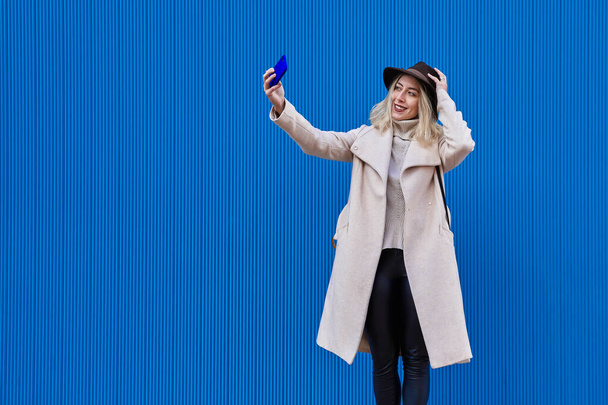 Een mooie vrouw met een hoed die een zelfportret maakt met haar smartphone op een blauwe achtergrond - Foto, afbeelding