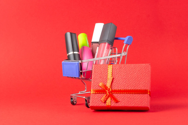 A set of lipstick cosmetics and lip glosses, powder, eyeshadow and a shopping trolley on a bright red background. The concept of buying cosmetics, online shopping, holiday - Foto, Imagem