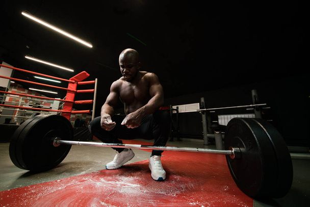 A man heavyweight african american smears his palms with magnesium or talc and prepares to lift the bar in the gym - Foto, afbeelding