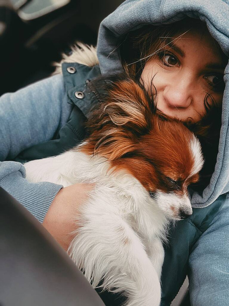 Uma jovem se senta em um carro com uma jaqueta azul e um capuz e segura um cachorro dormindo em seus braços.  - Foto, Imagem