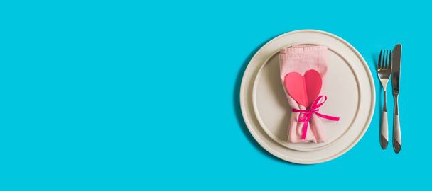 Banner with served table with plate and cutlery for the celebration of Valentine's Day. On the plate is napkin with paper heart. Flatlay website banner on bright blue background. Top view - 写真・画像