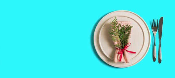 Banner with served table with empty plate and cutlery for celebration of Christmas and New Year. On plate is napkin with Christmas tree branch. Flatlay banner on bright blue background. Top view - Foto, Imagen