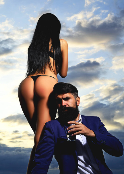 Bearded businessman in elegant suit with glass of whiskey. Rich man drink. Sencual couple in love. Womans butt in bikini. - Photo, image