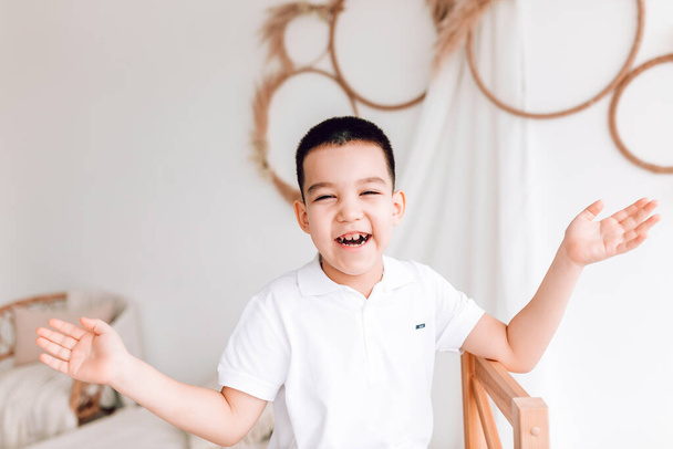 Ένα μικρό χαρούμενο αγόρι με λευκό μπλουζάκι χαμογελάει με τα χέρια απλωμένα. Ημέρα των παιδιών, η έννοια της χαράς των παιδιών - Φωτογραφία, εικόνα