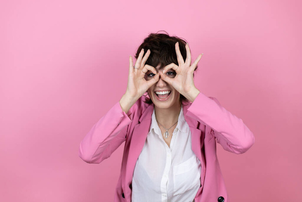 Молодая деловая женщина на изолированном розовом фоне делает хорошо жест шокирован улыбающимся лицом, глаза смотрят сквозь пальцы - Фото, изображение