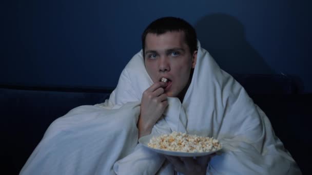 Shocked handsome caucasian Man watches TV movie with blanket in evening and eats popcorn on sofa at home, he is very interested in what he sees on screen and cannot take his eyes off. Close-up - Materiał filmowy, wideo