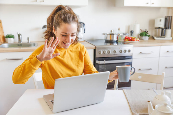 Uśmiechnięta dziewczyna macha ręcznym wideo dzwoniąc do rodziny przez kamerę internetową. Kobieta z laptopem o wirtualnym spotkaniu wideokonferencji rozmowy siedzi w kuchni w domu. Nowa normalna samoizolacja na odległość - Zdjęcie, obraz