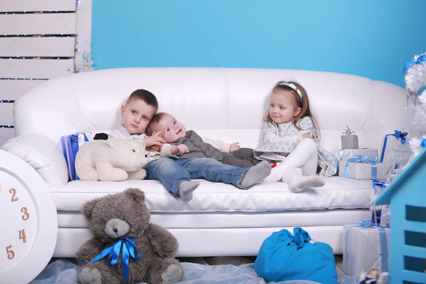 Δύο μικρά αγόρια και κορίτσια κάθονται σε λευκό καναπέ κοντά σε χριστουγεννιάτικα δώρα. Πρωτοχρονιάτικες διακοπές. Οικογένεια, ευτυχία, διακοπές, Χριστούγεννα έννοια. - Φωτογραφία, εικόνα
