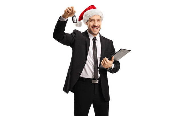 Smiling man in a suit showing a car key and wearing a santa claus hat isolated on white background - Фото, изображение