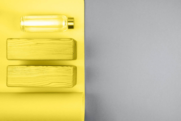 黄色のヨガマット、グレーの背景に2つの木製ブロックとエコ再利用可能なガラス水ボトルのトップビュー。テキスト用のスペース。流行色2021年. - 写真・画像