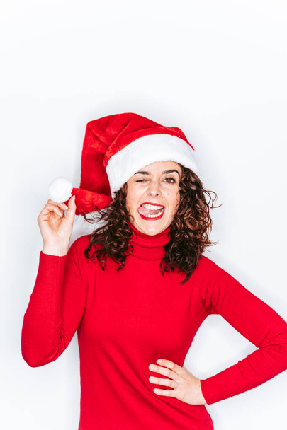 Un divertente ritratto di donna con un cappello da Babbo Natale. Lei indossa un maglione rosso e sta guardando la fotocamera con la lingua fuori. Concetto ritratto Natale. - Foto, immagini