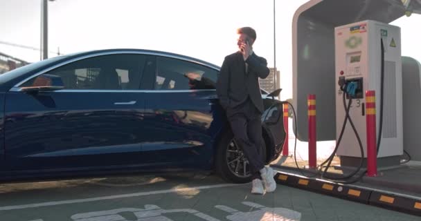 Caucasian businessman using a smart phone and waiting for the power supply to connect to electric cars to charge the battery in the car. Connect the charger of the electric car from the charging - Footage, Video