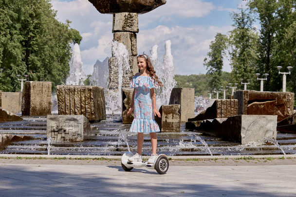 A beautiful girl in a blue dress rides a gyro scooter, hoverboard in summer. - Foto, Imagem