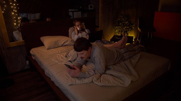 Женатая пара лежит на диване, девушка перекидывает ноги через парня и болтает по телефону, он занимается серфингом в социальных сетях - Фото, изображение