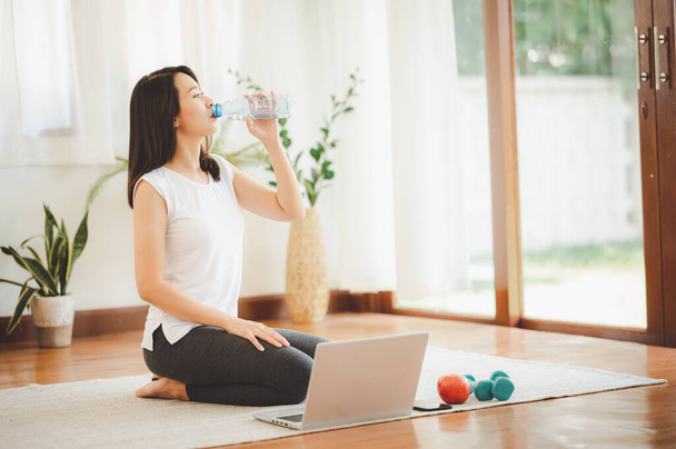 Healthy young Asian woman drinking from her water bottle while using laptop practicing yoga at home - Valokuva, kuva