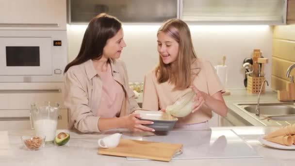 Images moyennes d'adolescente heureuse versant un mélange de crème glacée liquide dans un récipient spécial pour congeler debout à la table de cuisine avec maman - Séquence, vidéo