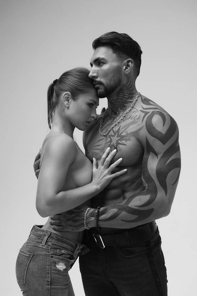 Zwart en wit zijaanzicht van topless vrouw en shirtloze man kijkend naar camera tegen grijze achtergrond - Foto, afbeelding