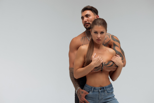 Strong shirtless male embracing and covering breast of attractive topless female on gray background - Photo, Image