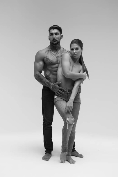 Black and white side view of topless woman and shirtless man looking at camera against gray background - Photo, Image