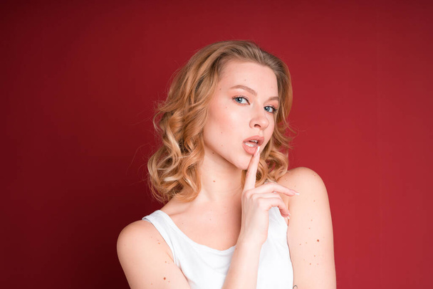Blond woman with curly hair teasing with her finger on mouth in white tank top isolated on red background. - Foto, imagen