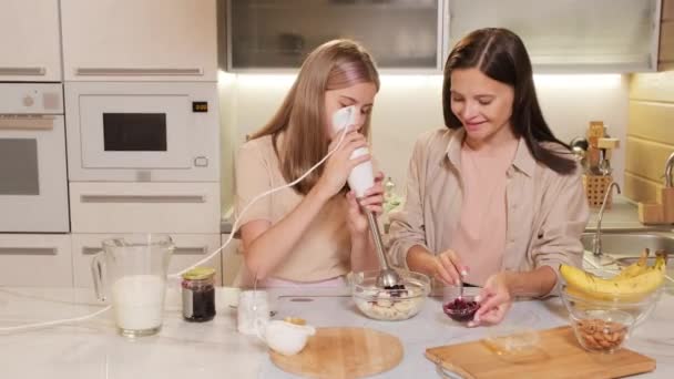 Tour de taille de jeune jolie adolescente mélangeant des ingrédients sucrés dans un bol pour crème glacée maison avec mélangeur d'immersion avec maman l'aidant - Séquence, vidéo