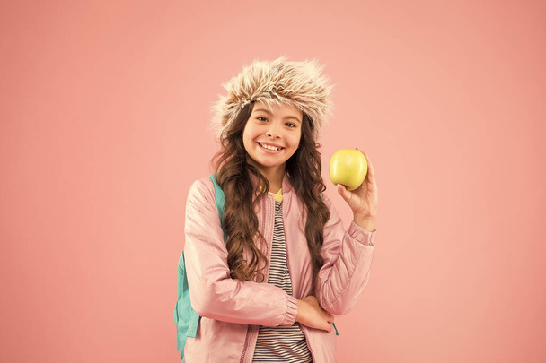 end of winter semester. holiday and vacation. kid pink backdrop. teen daily life. education and happy people schoolgirl with books after lesson. school break. small happy girl earflap hat eat apple - Foto, imagen