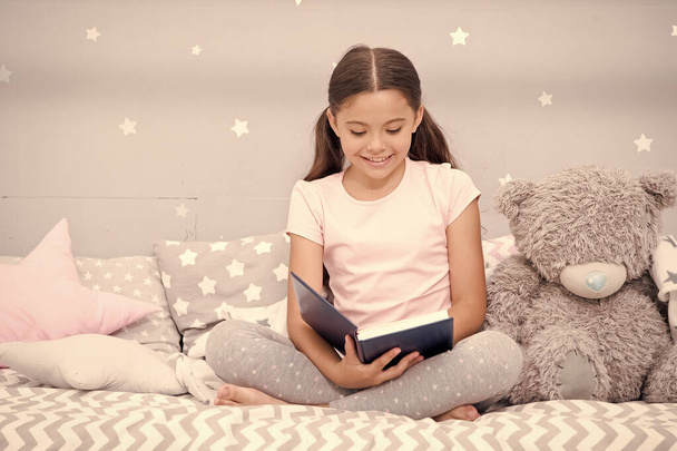 Dreaming big. Girl child in bed read book with teddy bear plush toy. Pleasant time in cozy bedroom. Girl kid cute pajamas relax and read book. Book about love. Favorite fairy tale. Literature hobby - Foto, afbeelding