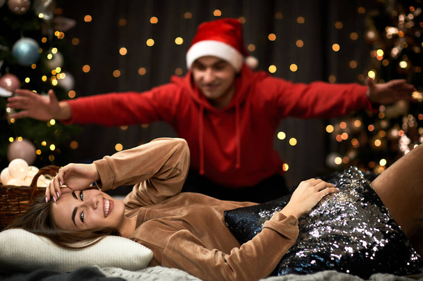woman is lying on bed and boy friend in a santa claus hat hid to surprise. - Zdjęcie, obraz