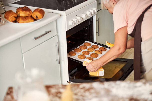 Aged lady getting baking sheet with cookies out of oven - Photo, Image